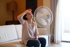 Woman sweating on a couch in front of a fan, indicating need for HVAC services during hot weather.
